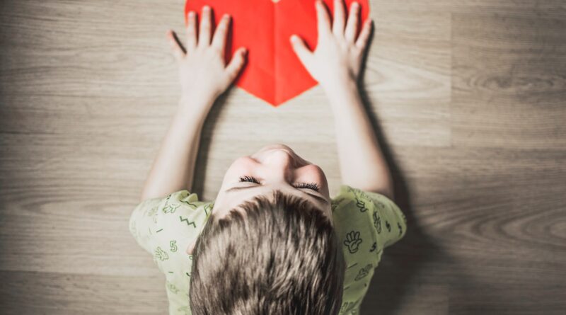 boy in green shirt holding red paper heart cutout on brown table